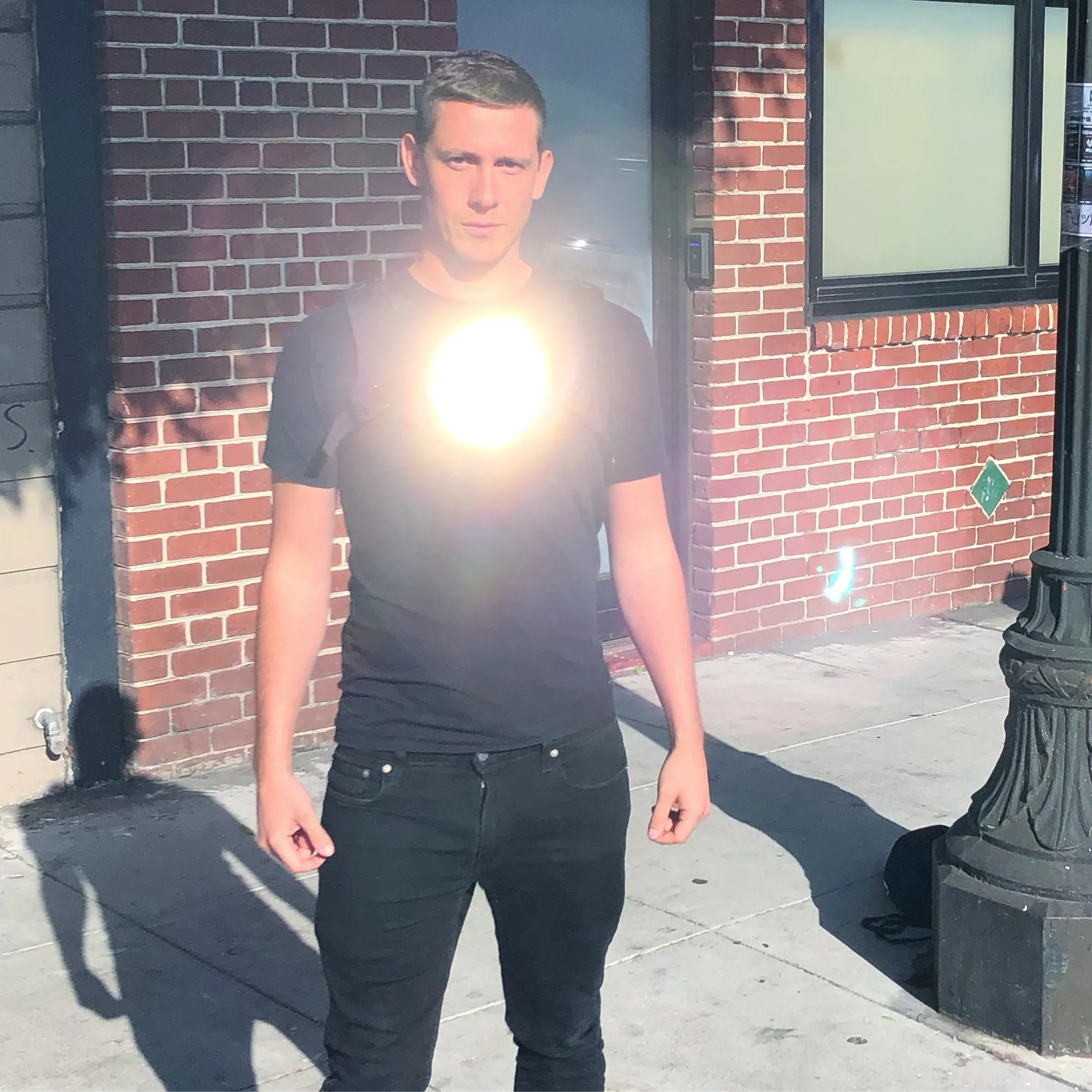 The author on a brick sidewalk in a black T-shirt with the Holovest at full brightness, a sphere of light at the chest flaring the lens